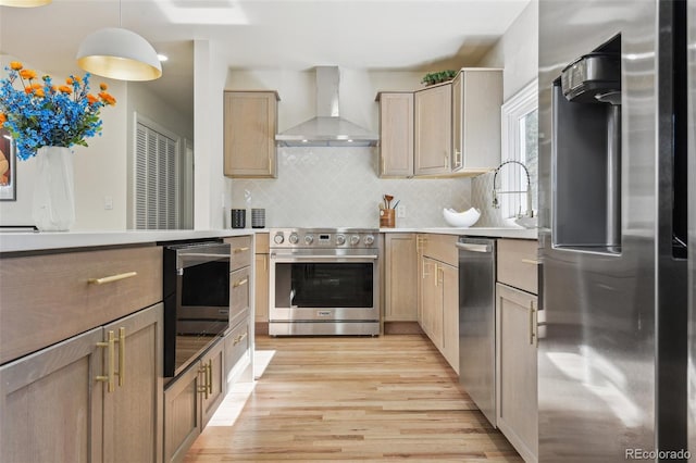 kitchen featuring hanging light fixtures, wall chimney exhaust hood, light wood finished floors, stainless steel appliances, and decorative backsplash