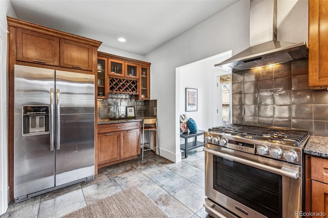 kitchen featuring appliances with stainless steel finishes, brown cabinets, decorative backsplash, and wall chimney exhaust hood