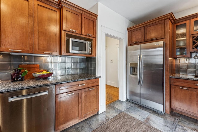 kitchen with stone tile floors, glass insert cabinets, brown cabinets, dark stone countertops, and stainless steel appliances