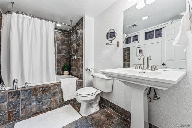 full bathroom with a textured wall, toilet, tiled shower / bath combo, visible vents, and baseboards