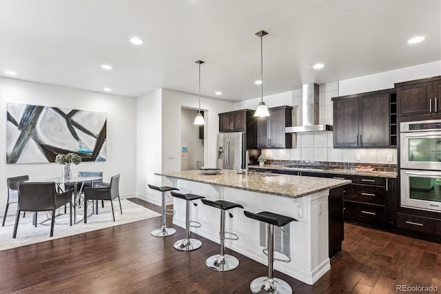 kitchen featuring appliances with stainless steel finishes, dark hardwood / wood-style flooring, wall chimney range hood, decorative light fixtures, and a center island with sink