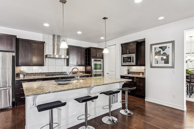 kitchen featuring dark brown cabinetry, a center island with sink, wall chimney exhaust hood, and stainless steel appliances