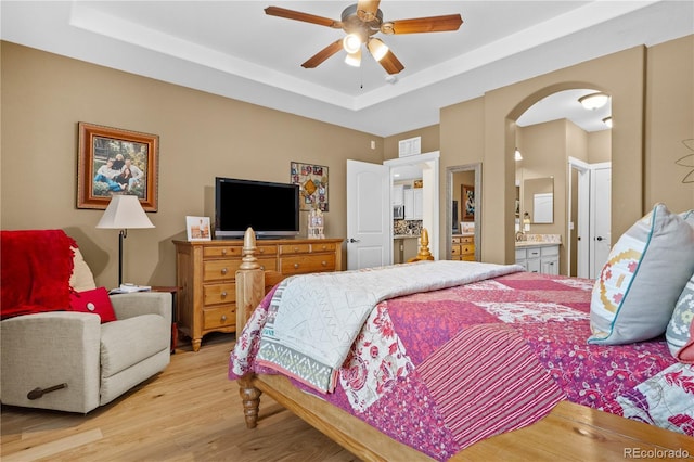 bedroom with a raised ceiling, light wood-type flooring, connected bathroom, and ceiling fan