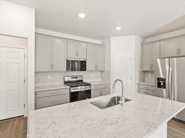 kitchen with appliances with stainless steel finishes, sink, a kitchen island with sink, light stone counters, and light hardwood / wood-style flooring