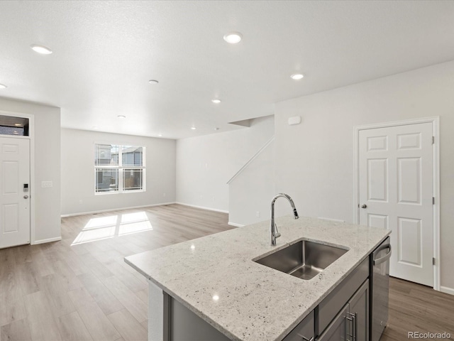 kitchen featuring an island with sink, sink, stainless steel dishwasher, light stone countertops, and light wood-type flooring