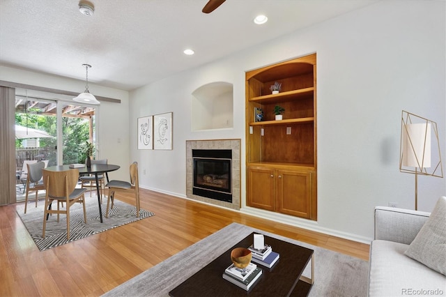 living room with a textured ceiling, a tile fireplace, built in features, ceiling fan, and light hardwood / wood-style floors