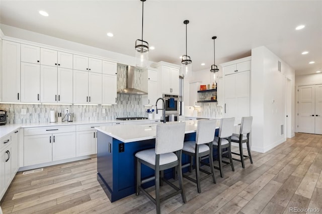 kitchen with wall chimney range hood, a center island with sink, a kitchen bar, white cabinetry, and hanging light fixtures