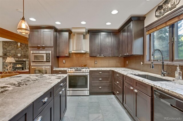 kitchen featuring stainless steel appliances, hanging light fixtures, decorative backsplash, wall chimney exhaust hood, and a stone fireplace