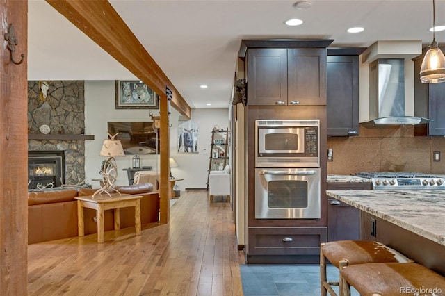 kitchen featuring light hardwood / wood-style flooring, wall chimney range hood, light stone counters, stainless steel appliances, and a fireplace