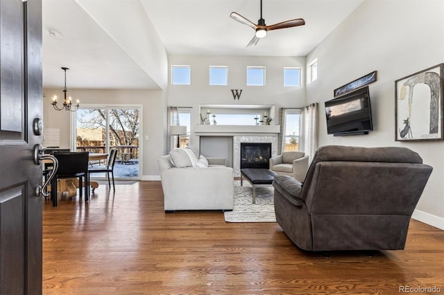 living room featuring a high ceiling, a tiled fireplace, wood finished floors, baseboards, and ceiling fan with notable chandelier