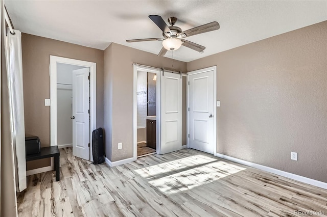 unfurnished bedroom with connected bathroom, light hardwood / wood-style flooring, ceiling fan, and a barn door