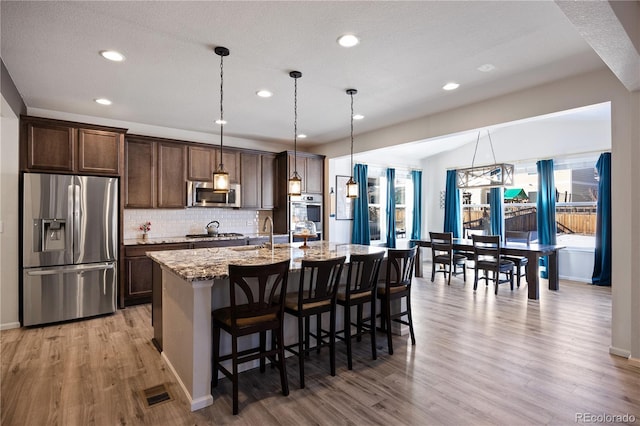 kitchen featuring appliances with stainless steel finishes, a kitchen island with sink, a kitchen breakfast bar, hanging light fixtures, and light stone counters