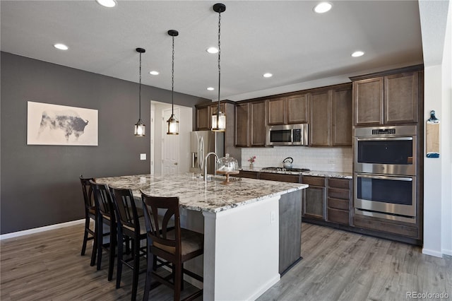 kitchen featuring decorative light fixtures, decorative backsplash, a kitchen island with sink, appliances with stainless steel finishes, and light stone counters