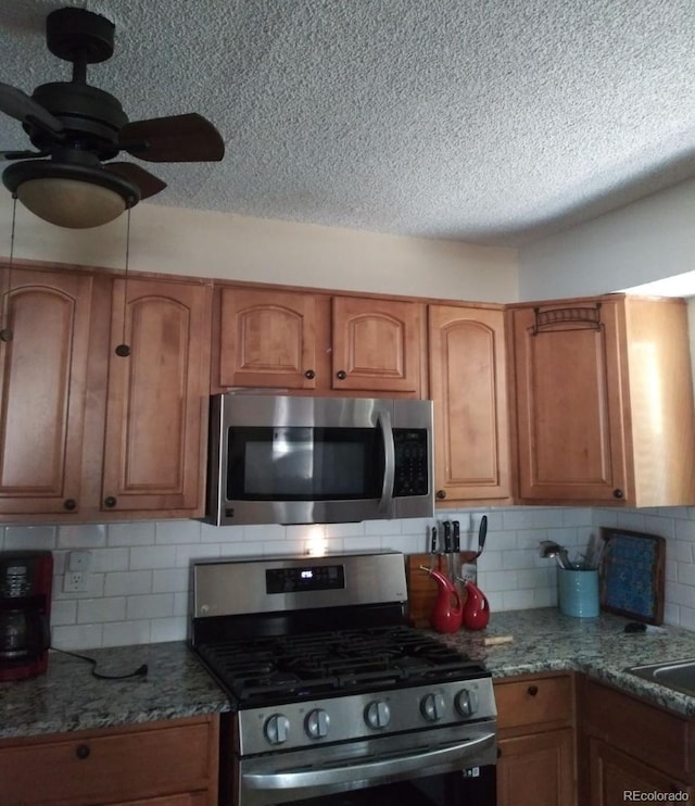 kitchen featuring stone counters, decorative backsplash, brown cabinetry, stainless steel appliances, and a ceiling fan