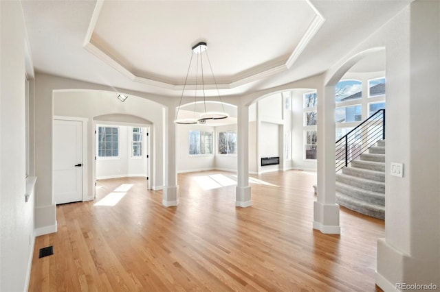 unfurnished dining area featuring light wood finished floors, a tray ceiling, arched walkways, stairs, and crown molding