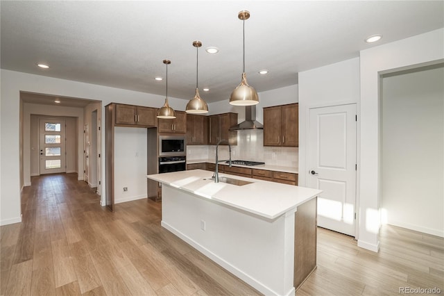 kitchen featuring appliances with stainless steel finishes, an island with sink, hanging light fixtures, light hardwood / wood-style flooring, and wall chimney range hood