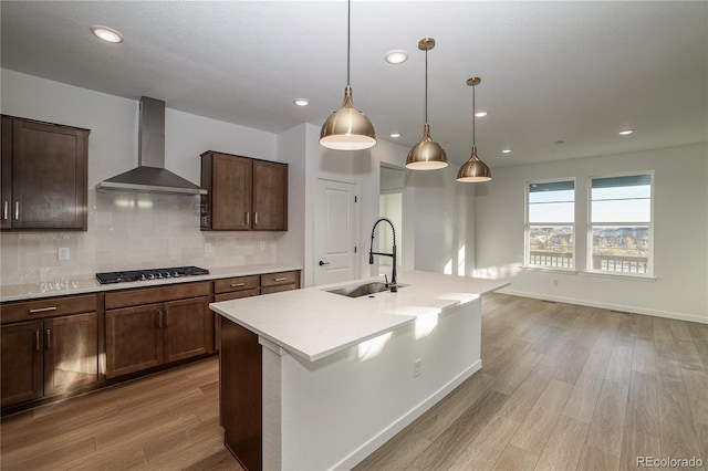 kitchen featuring sink, an island with sink, wall chimney range hood, light wood-type flooring, and decorative light fixtures