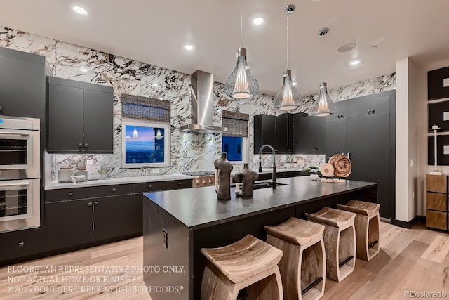 kitchen featuring double oven, hanging light fixtures, a kitchen island with sink, wall chimney range hood, and light hardwood / wood-style flooring