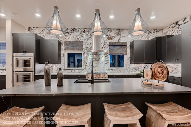 kitchen with a breakfast bar, double oven, decorative light fixtures, a large island with sink, and wall chimney exhaust hood