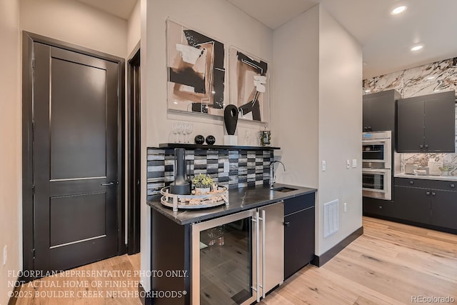 kitchen with tasteful backsplash, sink, beverage cooler, stainless steel double oven, and light wood-type flooring