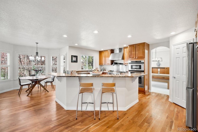 kitchen featuring wall chimney range hood, a textured ceiling, a chandelier, and appliances with stainless steel finishes