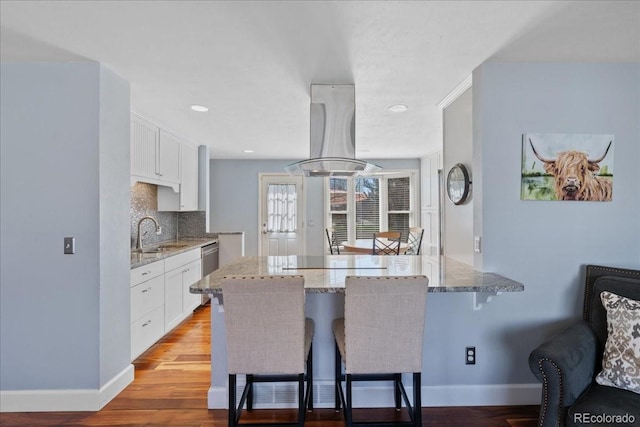 kitchen featuring a kitchen bar, island exhaust hood, light stone countertops, a sink, and white cabinetry