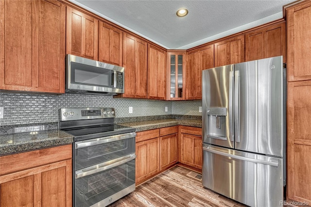 kitchen featuring glass insert cabinets, appliances with stainless steel finishes, brown cabinetry, backsplash, and light wood-style flooring