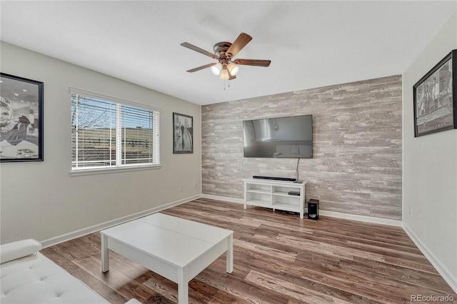 living area with baseboards, a ceiling fan, an accent wall, and wood finished floors
