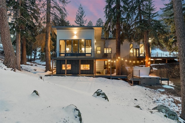 snow covered house featuring board and batten siding, a hot tub, a balcony, and stucco siding