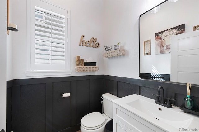 half bathroom featuring a decorative wall, toilet, vanity, and a wainscoted wall