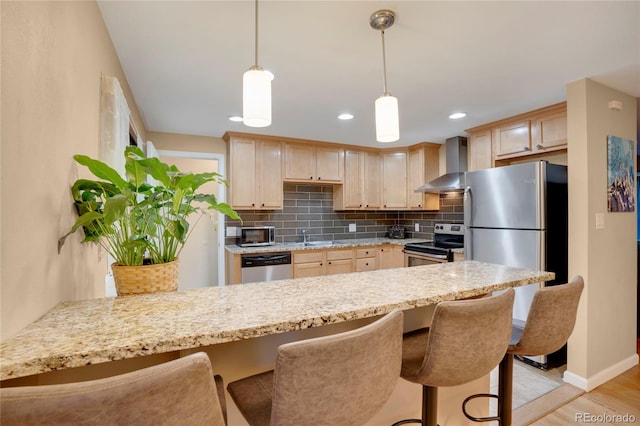 kitchen with light brown cabinets, a peninsula, wall chimney range hood, appliances with stainless steel finishes, and backsplash