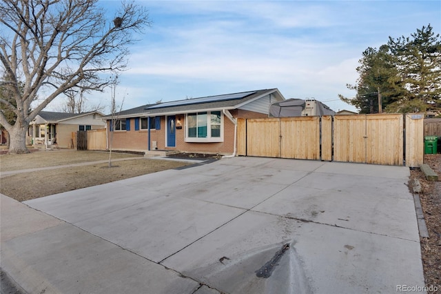 single story home featuring concrete driveway, a gate, fence, roof mounted solar panels, and brick siding