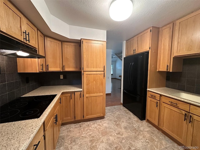 kitchen featuring brown cabinets, backsplash, a textured ceiling, under cabinet range hood, and black appliances