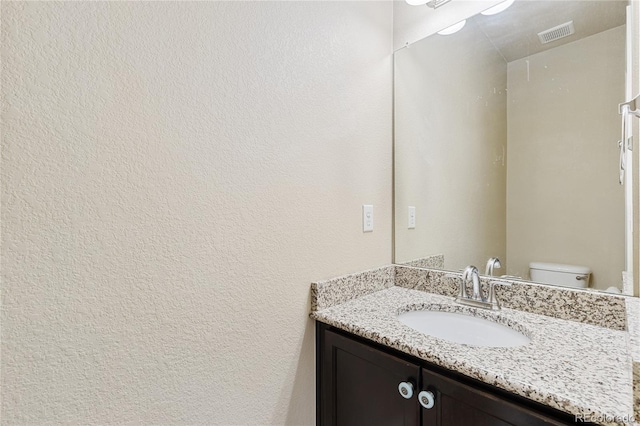 half bath with a textured wall, vanity, visible vents, and toilet