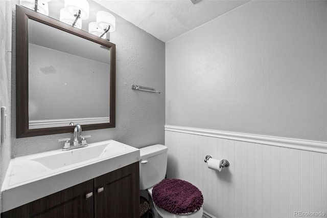 half bath featuring a textured wall, toilet, a wainscoted wall, a textured ceiling, and vanity