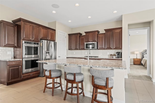 kitchen featuring a kitchen breakfast bar, a kitchen island with sink, appliances with stainless steel finishes, light stone counters, and tasteful backsplash