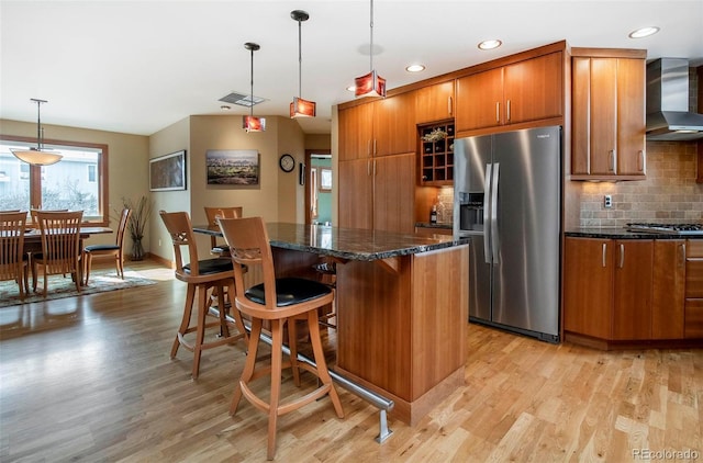 kitchen featuring stainless steel appliances, wall chimney range hood, light wood-type flooring, a center island, and brown cabinetry