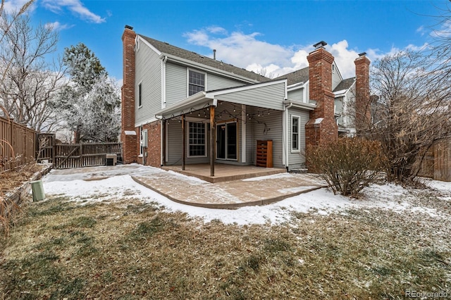 snow covered house featuring a patio area, a fenced backyard, brick siding, and a chimney