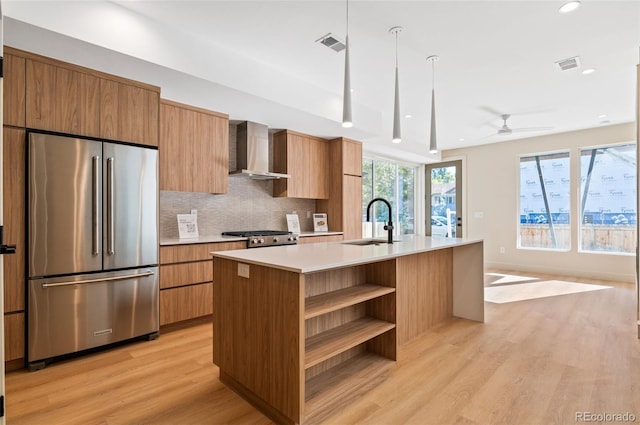 kitchen featuring appliances with stainless steel finishes, light wood-type flooring, backsplash, an island with sink, and wall chimney exhaust hood