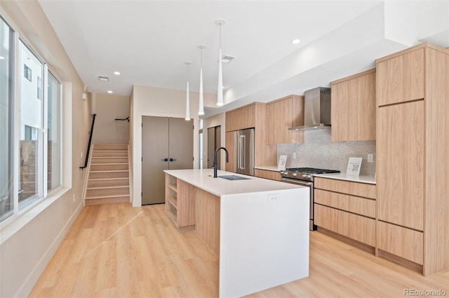 kitchen with wall chimney exhaust hood, an island with sink, light hardwood / wood-style floors, sink, and stainless steel range with gas stovetop