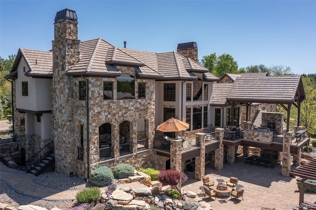 rear view of house featuring a chimney, stone siding, a patio area, and an outdoor living space with a fireplace