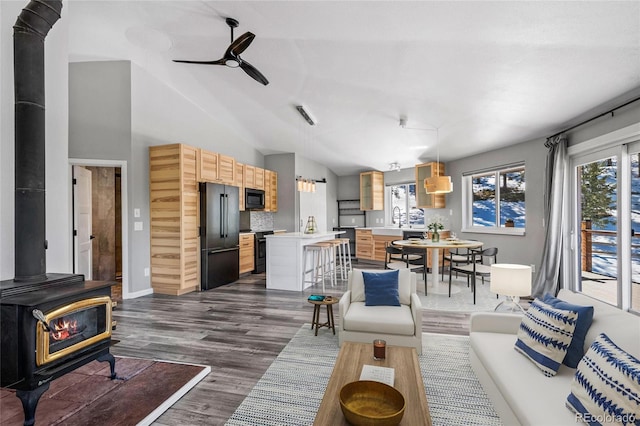 living room featuring vaulted ceiling, dark wood-style flooring, a wood stove, and a ceiling fan