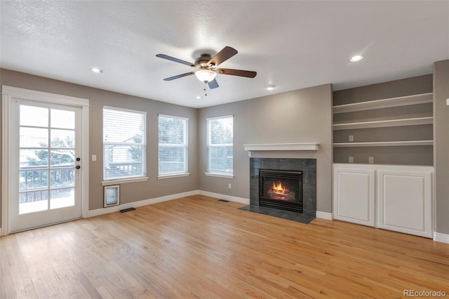 unfurnished living room with ceiling fan, a fireplace, a textured ceiling, built in shelves, and light wood-type flooring