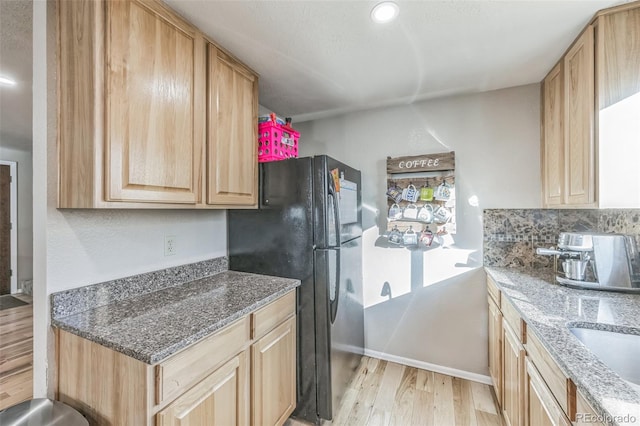 kitchen with backsplash, stone counters, black fridge, light wood-type flooring, and light brown cabinetry