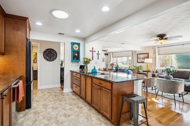 kitchen with a kitchen bar, black fridge, ceiling fan, light hardwood / wood-style flooring, and dark stone countertops