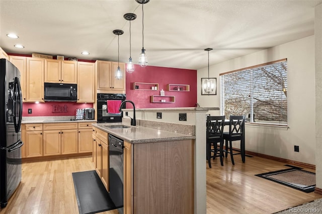 kitchen featuring light wood-type flooring, black appliances, light brown cabinets, decorative light fixtures, and a center island with sink