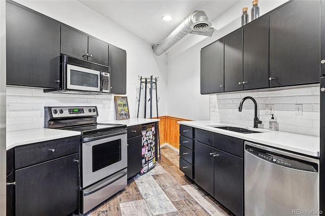 kitchen with appliances with stainless steel finishes, a sink, light wood finished floors, dark cabinetry, and wainscoting