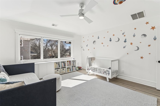 bedroom with wood finished floors, visible vents, baseboards, ceiling fan, and a nursery area