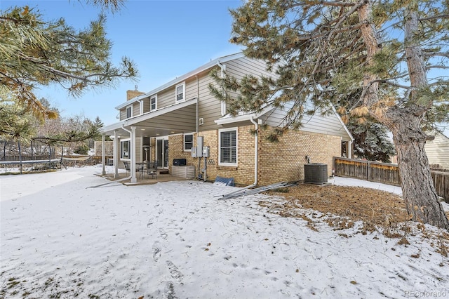 snow covered rear of property featuring a trampoline, brick siding, a chimney, fence, and cooling unit