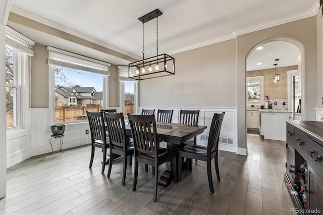 dining area featuring arched walkways, dark wood-style flooring, a wainscoted wall, a decorative wall, and ornamental molding